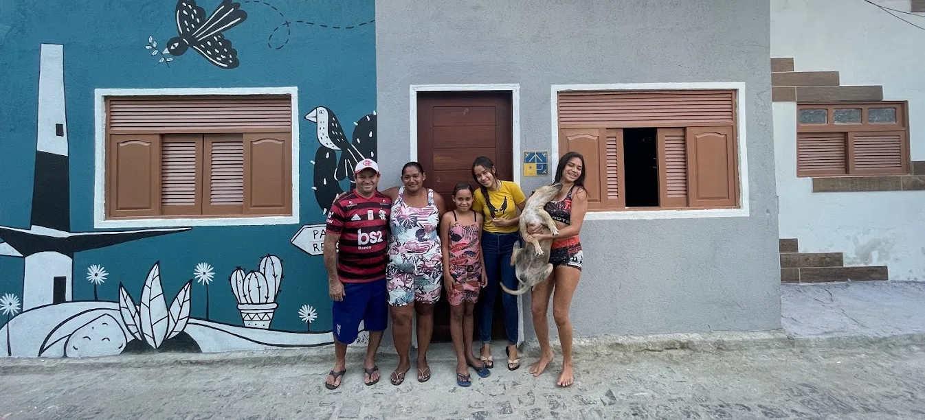 Família reunida diante da casa reformada pela ReforAMAR, com a fachada cinza, a porta de madeira e o mural pintado ao lado