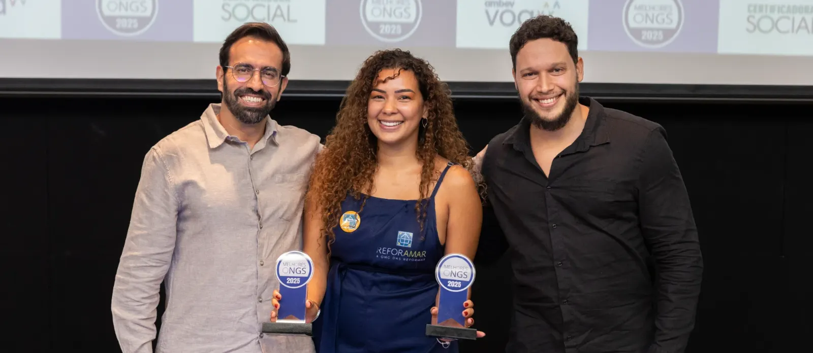 Representante da ReforAMAR sorrindo, entre dois homens, segurando os dois troféus do Prêmio Melhores ONGs 2025