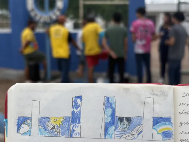 Caderno com o desenho da pintura artística planejada para a fachada de uma instituição, com o grupo do projeto ao fundo